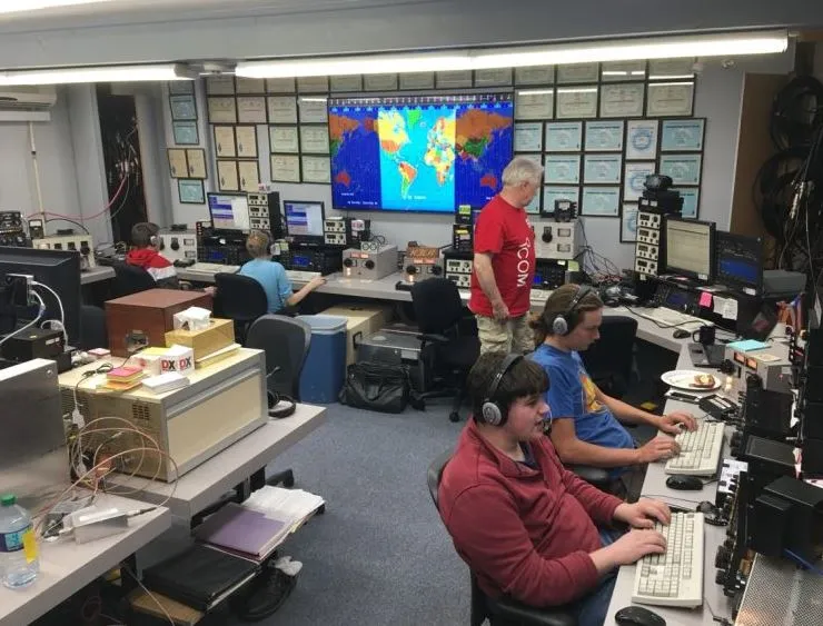 Operators wearing headphones working side by side at stacked radios in a multi operator contest station, with a world map and walls of awards behind them, during a ham radio contest weekend